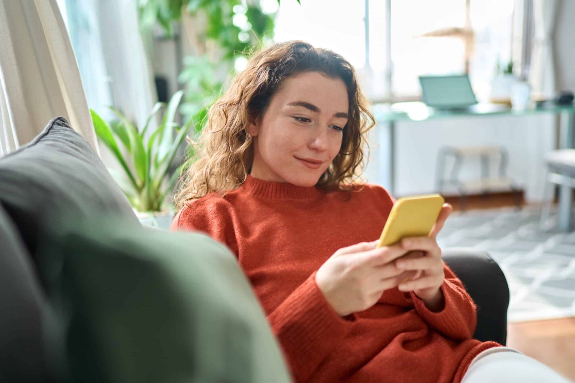Smiling relaxed young woman sitting on couch using mobile phone technology.